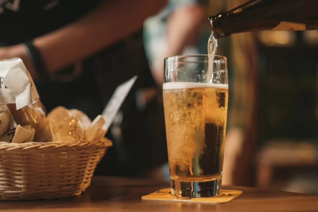 Bottle of beer being poured into a pint glass, with a basket of food behind it